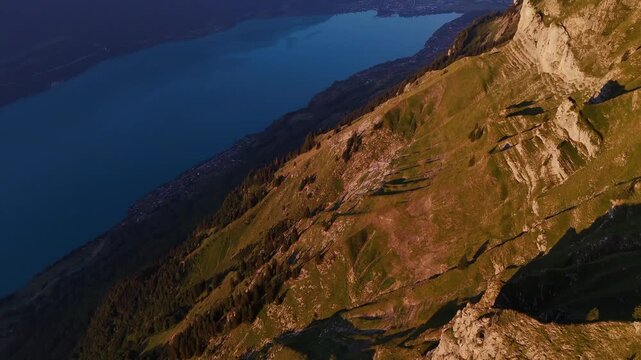 Drone view of sunlit alpine slopes of Augstmatthorn descending toward deep blue Lake Brienz in the Swiss Alps at sunrise.