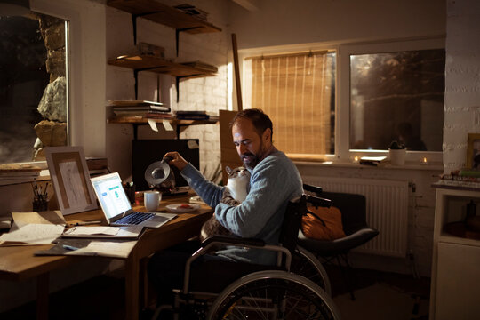 Man in wheelchair working from home office with cat and coffee at night
