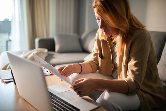 Focused woman reviewing paperwork on laptop at home