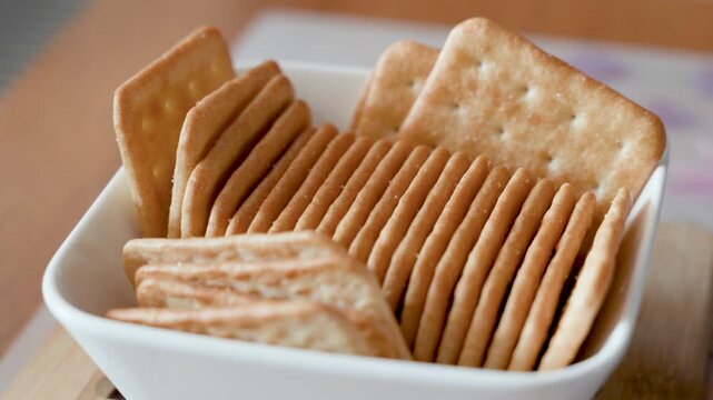Crispy golden crackers arranged in white ceramic bowl on wooden table, salty snack close up