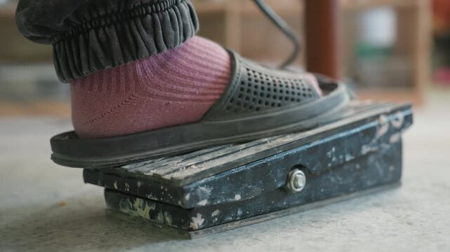 Pottery pedal foot action in studio. Apprentice modulates wheel speed with worn metal pedal, pink sock and slipper visible, soft daylight in workshop, unseen hands shaping clay while student practices