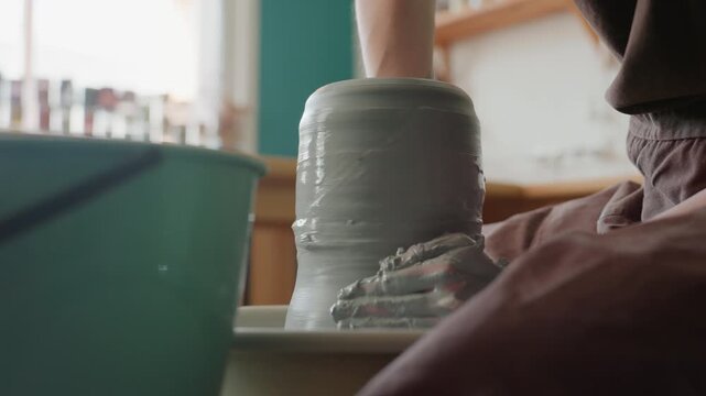 Hands forming tall clay vessel on wheel, ceramic artist in atmospheric workshop shaping vase, steady upward pulls and wet fingers, green bucket and soft daylight, closeup of texture and motion