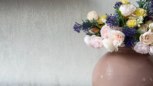 Elegant pastel artificial flower arrangement in a glossy pink vase against a neutral textured wall, decorative floral interior composition with copy space.