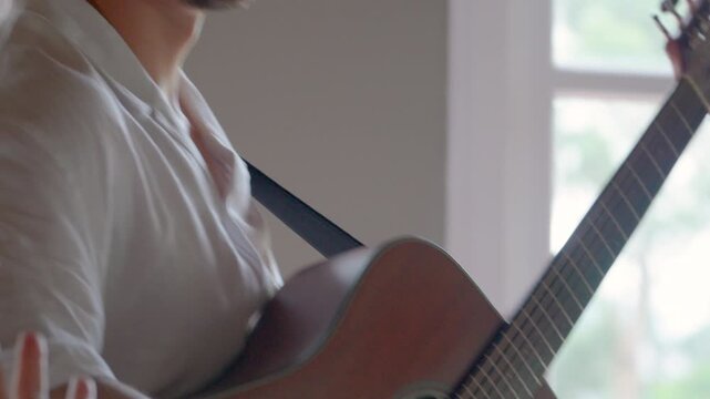 Musician strumming a wooden acoustic guitar in a brightly lit room next to a window.