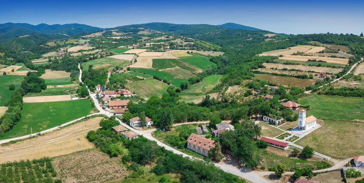 Aerial view capturing a peaceful rural village with houses, church, roads, and agricultural fields surrounded by rolling green hills