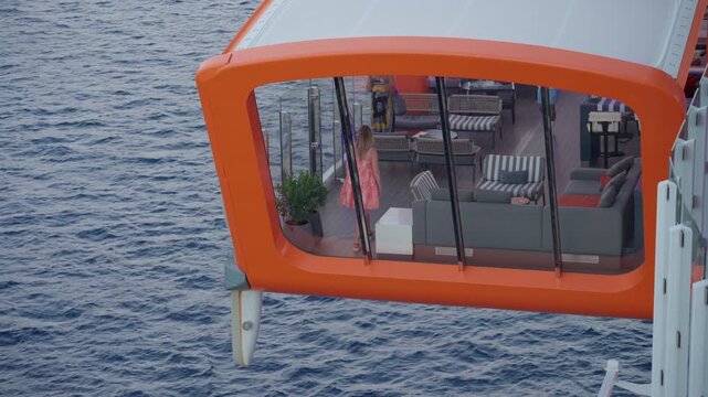 Cruise liner observation lounge extends over Caribbean Sea as woman in pink dress walks past modern sofas and striped cushions with deep blue ocean waves visible through panoramic glass windows.