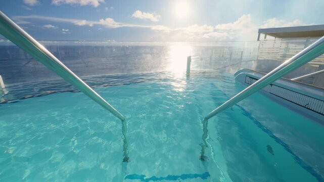 Cruise liner infinity pool sparkles with crystal clear turquoise water as sunlight reflects on the surface. Glass barrier reveals the Caribbean Sea horizon with white clouds and blue sky beyond.