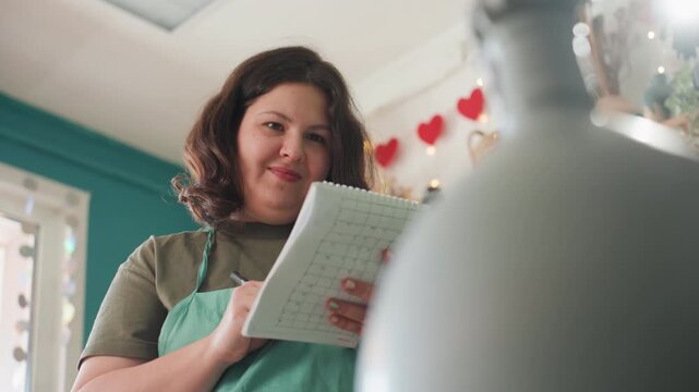 Studio owner preparing pottery inventory list checking vases and prices in notebook, green apron, vase foreground, natural window light, heart garland, product display and packaging, shipping prep,