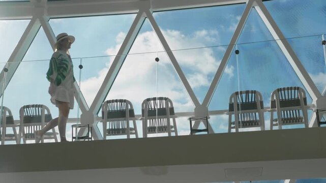Woman in straw hat and green tropical shirt walks along modern cruise ship deck featuring geometric glass ceiling and lounge chairs with blue Caribbean Sea sky visible through triangular windows.
