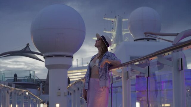 Young woman wearing bikini top and sun hat stands at glass railing on cruise liner deck during twilight. Satellite domes and ship structures visible against dramatic cloudy sky in Caribbean Sea.