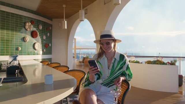 Stylish woman wearing straw hat and green striped blouse relaxes at Mediterranean-style outdoor bar with white arched columns on cruise ship deck.