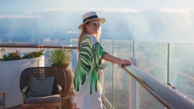Stylish woman in straw hat and tropical print blouse stands on luxury cruise liner deck in Caribbean Sea. She smiles while gazing at ocean horizon during golden sunset with glass railings and plants.