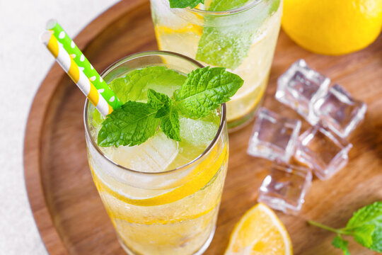 Close up of refreshing homemade lemonade with fresh lemon slices, mint leaves, ice cubes with soda water and paper straw served in glass with water droplets on wooden kitchen counter background.