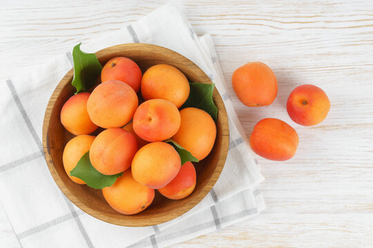 Top view of ripe apricots with green leaves arranged in wooden bowl on on white tile background. Fresh seasonal harvest, summer fruit, healthy eating, dessert, food ingredient