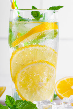 Close up, macro of refreshing homemade lemonade served in clear glass with fresh lemon slices, mint leaves, ice cubes and paper straws on a light kitchen counter background