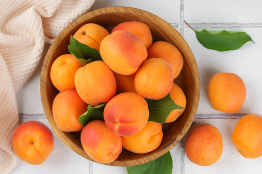 Top view of ripe apricots with green leaves arranged in wooden bowl on on white tile background. Fresh seasonal harvest, summer fruit, healthy eating, dessert, food ingredient