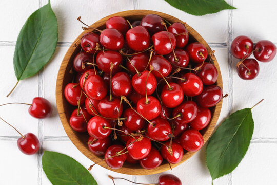 Top view of fresh ripe red sweet cherries in wooden bowl with green leaves on white tile textured background. Organic summer fruit, juicy berries, natural freshness, healthy eating, seasonal food