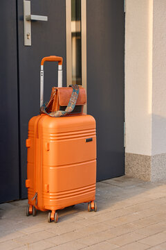 Orange rolling suitcase with shoulder bag stands beside a front door