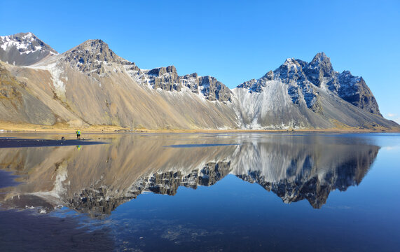 Vestrahorn mountain stokksnes reflecting on still water lagoon with two people walking on the black sand beach. Iceland Icelandic landmark landscape