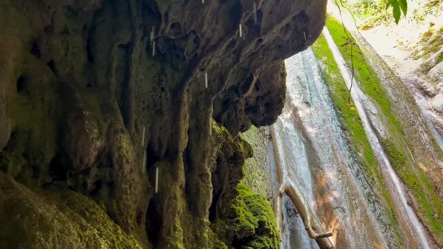 Water drips from the stone vault of the cave against the background of a waterfall flowing down the mossy rocks. Concept: mysterious cave, forest waterfall, unity with nature, hiking, the power of the
