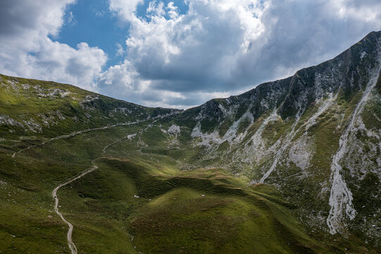 Aerial view of alpine cirque with winding hiking trail in the Bernese Oberland - green pastures under dramatic clouds