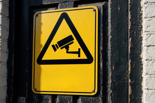 Yellow square sign with black border warns of surveillance camera. Symbol advises caution and security. Sign mounted on weathered dark wood next to brick wall.