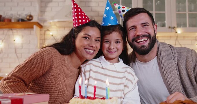 Family birthday with cake and candles at home. Smiling happy parents and child daughter in party hats sit near a cake in a cozy festive kitchen with gifts. Celebration and holiday concept. 