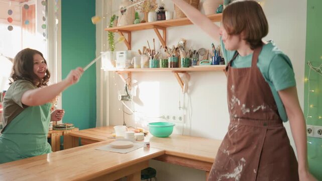 Pottery workshop playful tool duel between two artisans, tapping brushes and sponges over counter. Lively motion, rhythmic exchange and bright window light, friendly competition and shared laughter