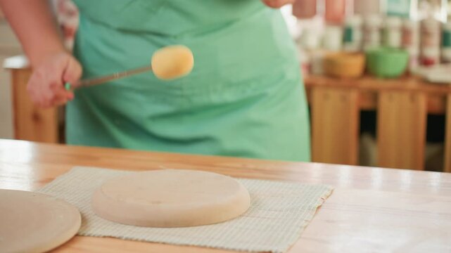 Ceramicist pottery workshop sponging bisque slab, closeup hands holding sponge tool and smoothing surface with steady rhythmic strokes, shelves of glazes and jars in background, green apron