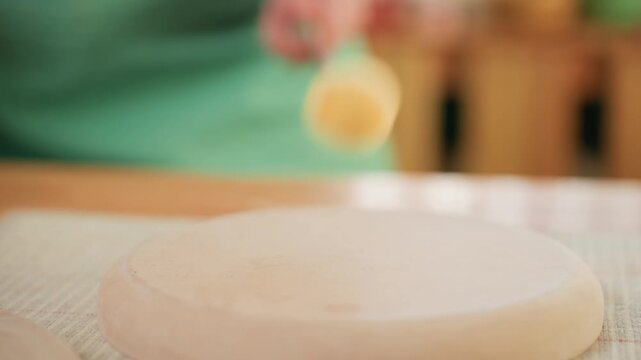 Closeup clay slab on workshop table, artisan smoothing disk with wooden tool, green apron in background, subtle beige texture and soft tactile finish, slow focused studio pottery process