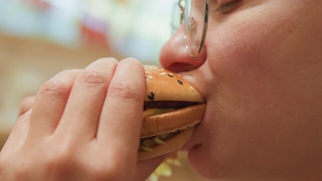 Woman eats burger. Closeup of woman biting cheeseburger. Candid shot of woman savoring savory burger with relish. Close portrait of woman biting into cheeseburger with messy sauce and glasses