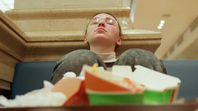 Takeout boxes foreground reflective young person seated against stone planter, head tilted upward, glasses catching light, scattered wrappers and empty containers, quiet moment after shopping, moody