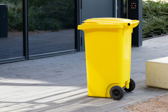 Yellow plastic waste bin with wheels positioned on a paved surface near a modern building, greenery visible in the background