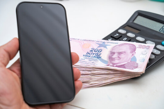Hand Holding Blank Smartphone Screen next to Stack of Turkish Lira Bills and Calculator