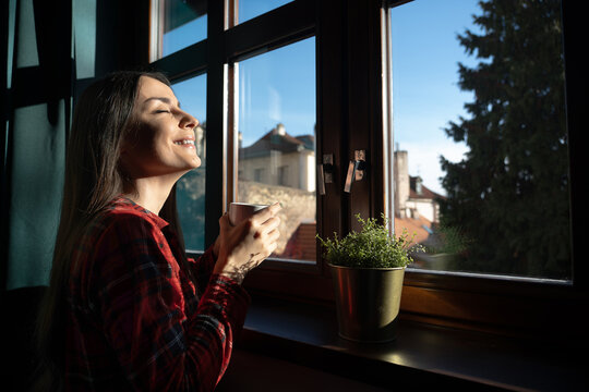 Smiling woman enjoying sunlight and coffee by window