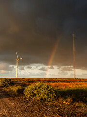 Obraz premium Stormy sky with rainbow arching over wind turbine and transmission tower. Open field with dry grass and shrubs under dramatic lighting. Blend of modern technology and nature scenery. Dark and moody.