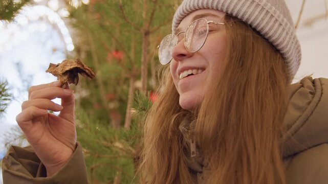 Young caucasian woman holding pinecone in warm holiday market glow, wearing beanie and glasses, smiling as she inspects texture and scent near twinkling lights and evergreen branches, candid closeup