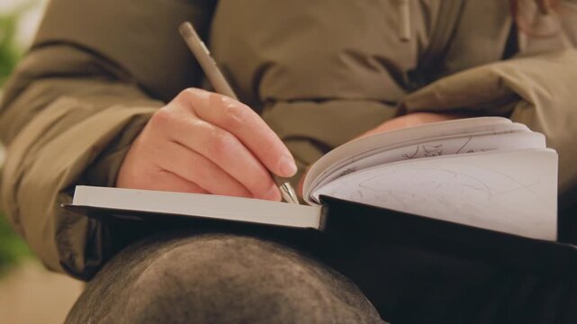 Closeup hands writing notebook outdoors, freelance writer journaling on knee in puffer jacket, ballpoint pen tracing lines on spiral pages, warm autumn tones, cozy focused mood, mindful planning