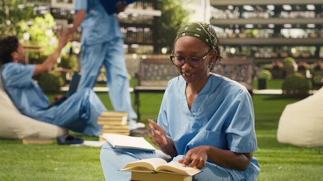 The medical student in blue scrubs reads and writes in a textbook during an outdoor study session, sitting on the grass with stacked books and classmates behind for medical school training.