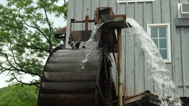 water wheel spins beside old barn and stream in mountain area during bright sunny day in summer season