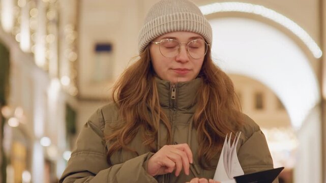 White woman in beanie checking planner, flipping pages and jotting notes with pen under glowing arch, calm organized mood as she balances study and holiday errands in lively evening market.