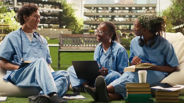 Medical students in scrubs stand together and review a laptop on a modern campus courtyard, discussing digital coursework and supporting collaborative healthcare learning, during peer tutoring.
