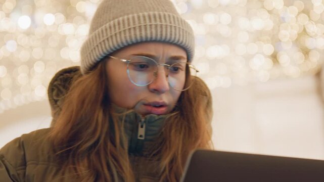 Young woman struggling with mobile payment at laptop, tense face under decorative lights, beanie and glasses, flips phone and makes support call while checking transaction status, holiday mall