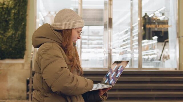 White woman outdoors on stairs working laptop, in urban shopping district, bundled in parka and beanie, red hair peeking, focused typing on slim laptop by illuminated boutique windows, evening bokeh