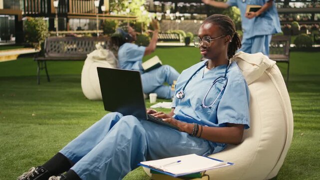 The woman medical student types on a laptop in blue scrubs with a stethoscope, taking notes while seated on a beanbag for outdoor medical study.