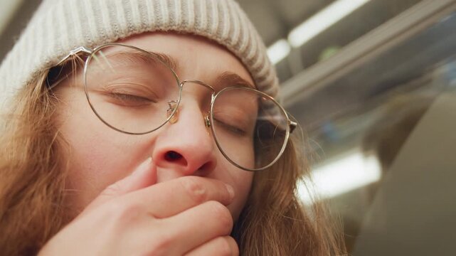 Tired morning commuter. Woman wearing beanie shows fatigue. Morning fatigue evident in woman with beanie and smile. Candid shot portraying drowsy woman with round glasses during early commute