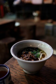 古い飲食店の木製テーブル。白碗に入った肉うどん。暗めの店内。