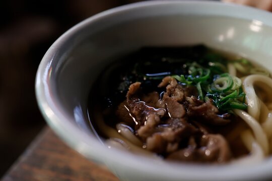 古い飲食店の木製テーブル。白碗に入った肉うどん。暗めの店内。