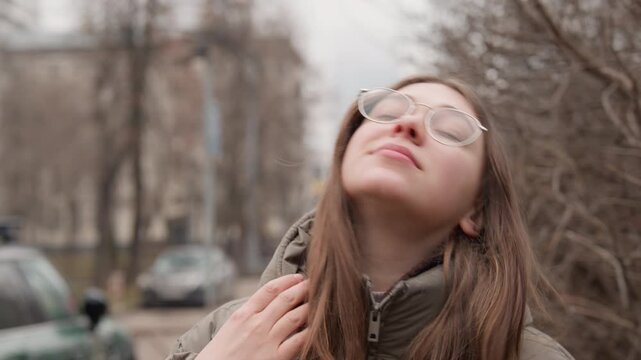 Young caucasian woman exhaling smoke on urban sidewalk, glasses fogging slightly, hood up and breath forming vapor in cold air, subtle smile and closed eyes between inhales, background traffic