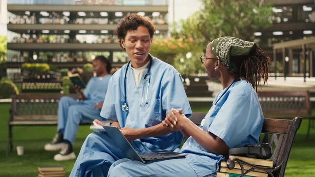 Diverse students in blue scrubs compare notes in an open textbook, discussing coursework during a peer tutoring session on campus bench, with academic materials supporting group study.